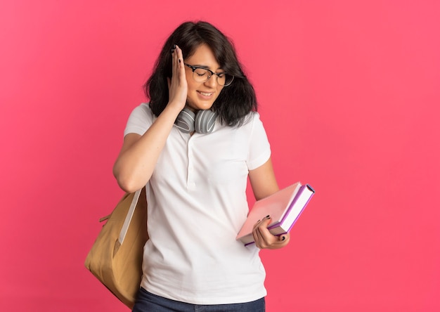 Joven colegiala bastante caucásica molesta con gafas, bolsa trasera y auriculares en el cuello cierra la oreja con la mano sosteniendo el libro y el cuaderno en rosa con espacio de copia