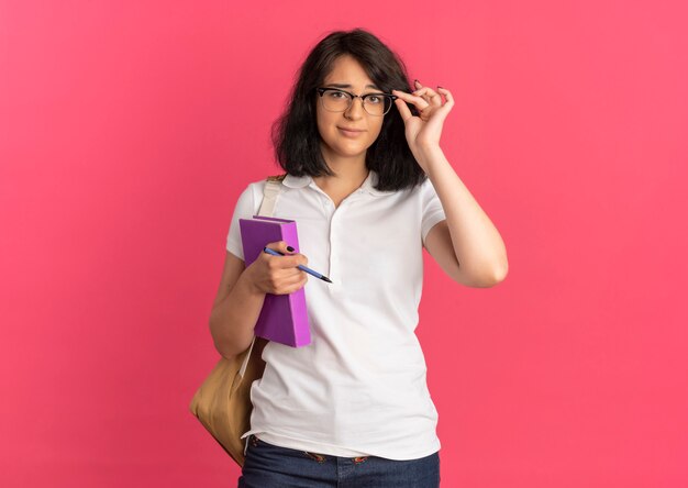 Joven colegiala bastante caucásica molesta vistiendo y sosteniendo gafas y bolsa trasera sostiene bolígrafo y libro en rosa con espacio de copia