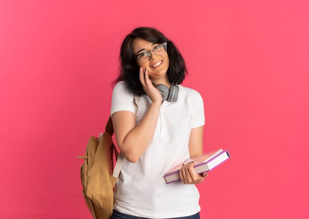 Joven colegiala bastante caucásica sonriente con gafas, bolsa trasera y auriculares en el cuello pone la mano en la cara sosteniendo el libro y el cuaderno en rosa con espacio de copia