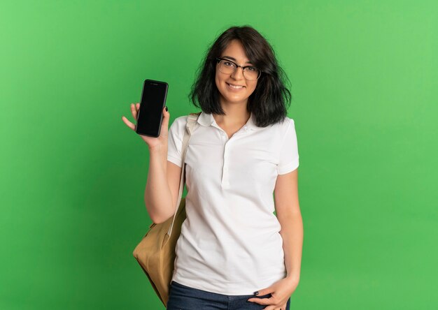 Joven colegiala bastante caucásica sonriente con gafas y bolsa trasera sostiene el teléfono en verde con espacio de copia