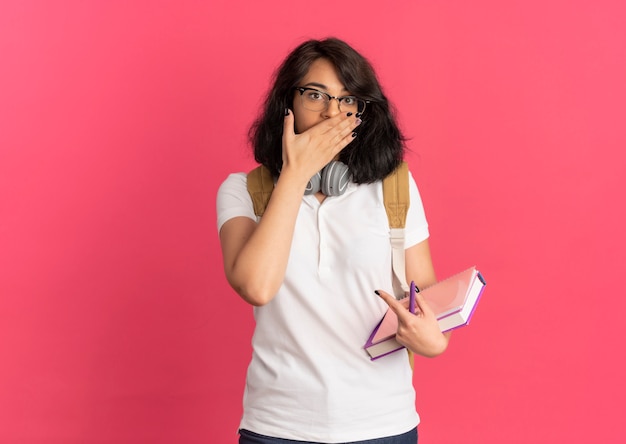 Joven colegiala bastante caucásica sorprendida con auriculares en el cuello con gafas y bolsa trasera pone la mano en la boca sosteniendo libros aislados en el espacio rosa con espacio de copia