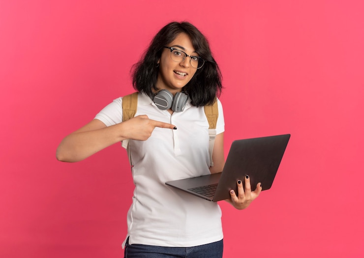 Foto gratuito joven colegiala bastante caucásica sorprendida con auriculares en el cuello con gafas y bolsa trasera sostiene y apunta a la computadora portátil en rosa con espacio de copia