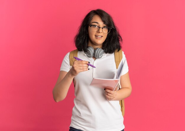 Joven colegiala bastante caucásica sorprendida con auriculares en el cuello con gafas y bolsa trasera sostiene bolígrafo y cuaderno en rosa con espacio de copia