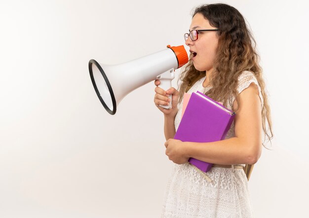 Joven colegiala bonita con gafas y bolsa trasera de pie en la vista de perfil sosteniendo libros hablando por altavoz aislado sobre fondo con espacio de copia