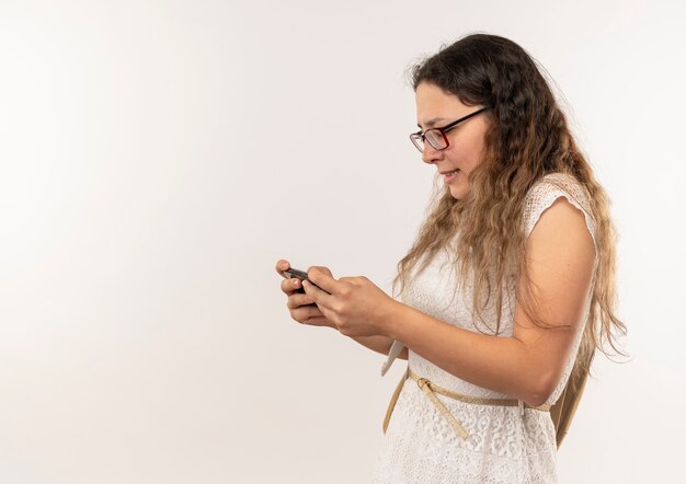 Joven colegiala bonita con gafas y bolsa trasera de pie en la vista de perfil usando su teléfono aislado sobre fondo con espacio de copia