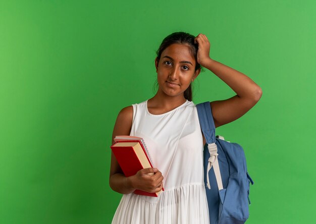 Joven colegiala con mochila sosteniendo el libro con el portátil y poniendo la mano en la cabeza.