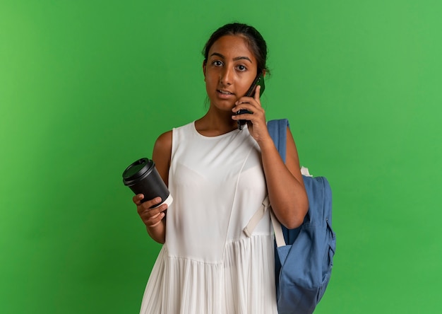 Joven colegiala con mochila sosteniendo una taza de café y habla por teléfono