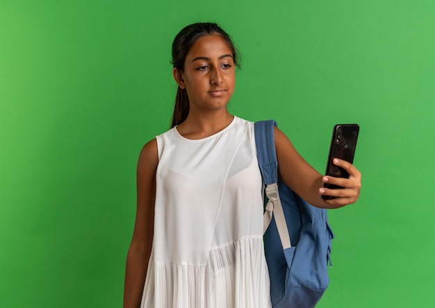 Joven colegiala vistiendo bolsa trasera sosteniendo y mirando el teléfono en verde