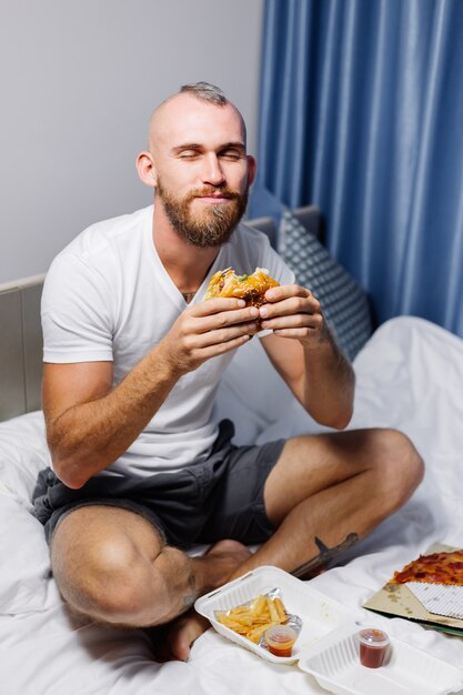 Joven con comida rápida en casa en el dormitorio en la cama