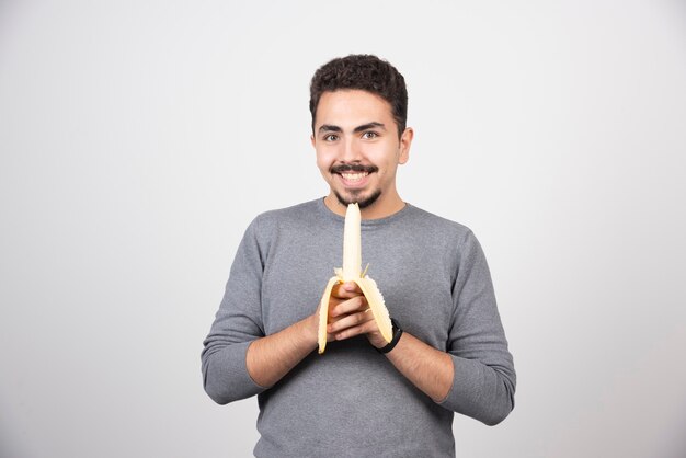 Un joven comiendo un plátano sobre una pared blanca.