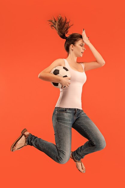 La joven como futbolista saltando y pateando la pelota en el estudio sobre un fondo rojo.