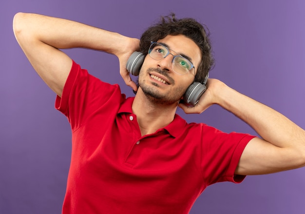 Joven complacido en camisa roja con gafas ópticas y con auriculares tiene las manos detrás de la cabeza aislada en la pared violeta