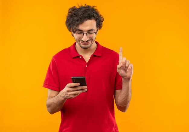 Joven complacido en camisa roja con gafas ópticas sostiene el teléfono y apunta hacia arriba aislado en la pared naranja