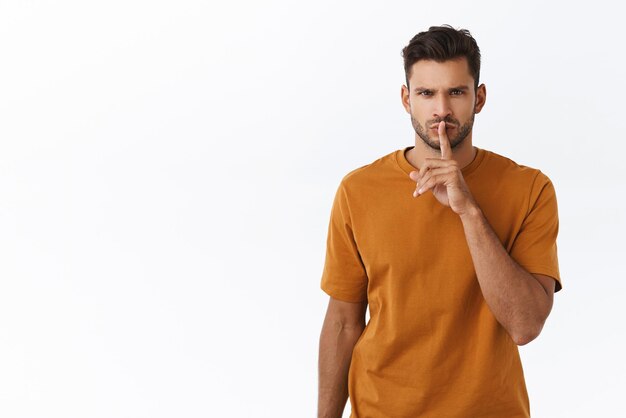 Un joven concentrado y estricto de aspecto serio con barba que se calla a la cámara con el dedo índice presionado contra los labios doblados frunciendo el ceño silenciando la demanda deja de hablar en voz alta di silencio por favor fondo blanco