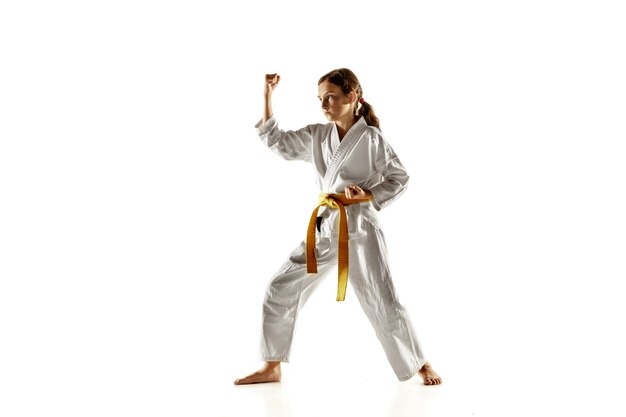 Joven confiado en kimono practicando combate cuerpo a cuerpo, artes marciales. Joven luchadora con entrenamiento de cinturón amarillo en la pared blanca. Concepto de estilo de vida saludable, deporte, acción.