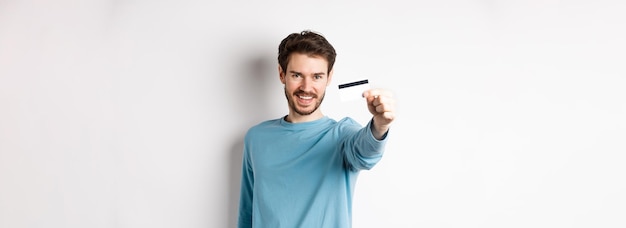 Foto gratuita un joven confiado sonriendo estira la mano y mostrando una tarjeta de crédito de plástico de pie en el respaldo blanco