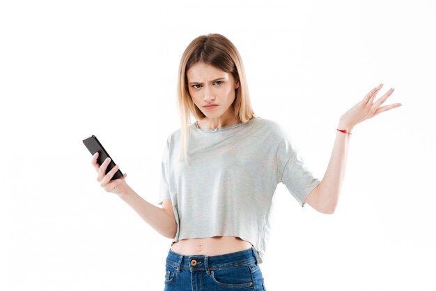 Joven confundida joven sosteniendo teléfono móvil y encogiéndose de hombros