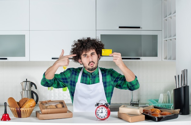 Joven confundido de pie detrás de la mesa varios pasteles y mostrando la tarjeta bancaria en la cocina blanca