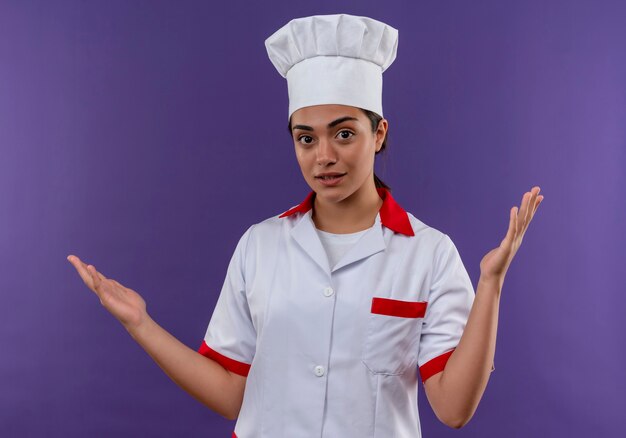 Joven confusa cocinera caucásica en uniforme de chef levanta las manos aisladas en la pared violeta con espacio de copia