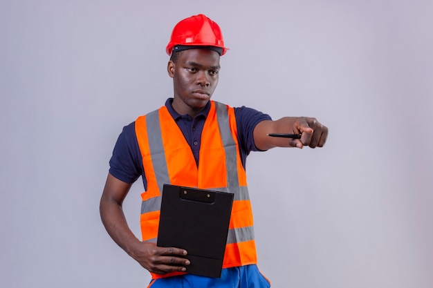 Joven constructor afroamericano hombre vestido con chaleco de construcción y casco de seguridad apuntando con el dedo índice a la distancia con expresión seria de pie