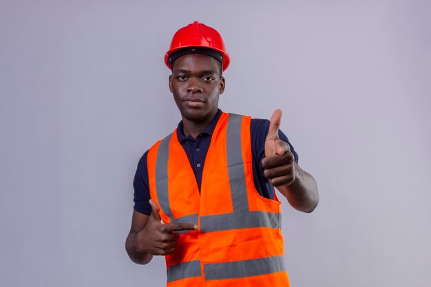 Joven constructor afroamericano hombre vestido con chaleco de construcción y casco de seguridad apuntando con el dedo mirando de pie confiado