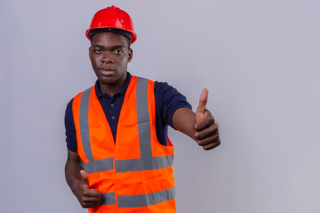 Joven constructor afroamericano hombre vestido con chaleco de construcción y casco de seguridad mostrando el pulgar de pie