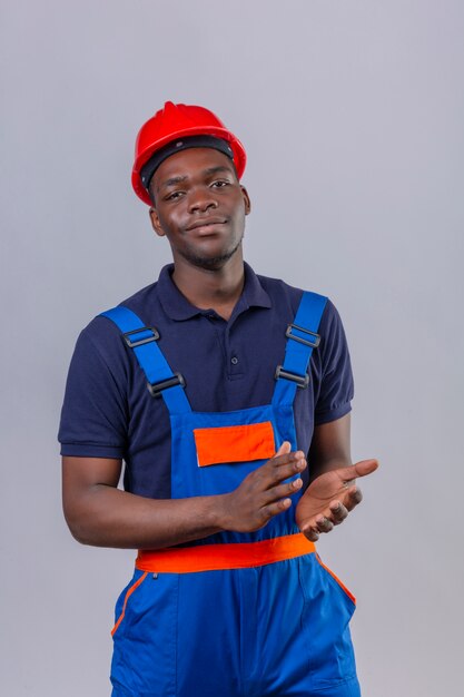 Joven constructor afroamericano hombre vestido con uniforme de construcción y casco de seguridad aplaudiendo con sonrisa de confianza en la cara de pie