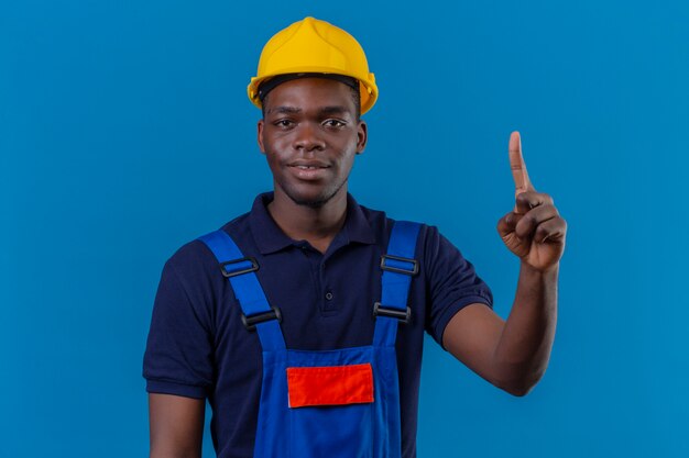 Joven constructor afroamericano hombre vestido con uniforme de construcción y casco de seguridad apuntando con el dedo hacia arriba con expresión de confianza de pie en azul