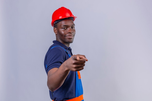 Joven constructor afroamericano hombre vestido con uniforme de construcción y casco de seguridad dedo acusador con sonrisa de confianza de pie