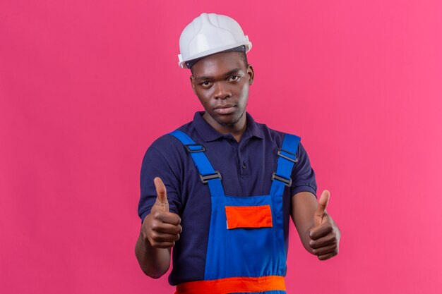 Joven constructor afroamericano hombre vestido con uniforme de construcción y casco de seguridad mostrando los pulgares para arriba en rosa