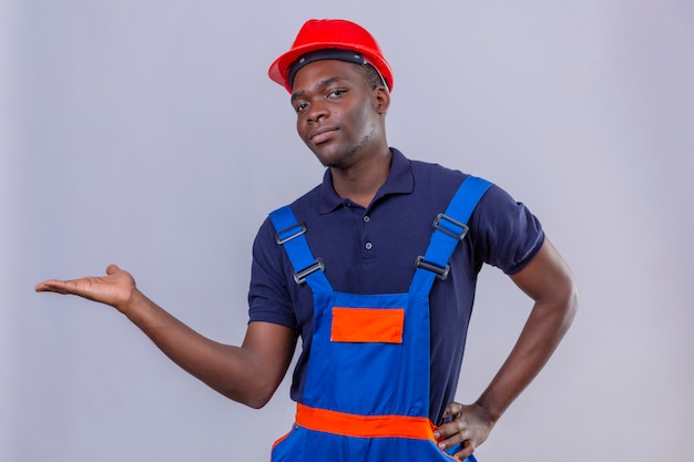 Joven constructor afroamericano hombre vestido con uniforme de construcción y casco de seguridad sonriendo amigable presentando y señalando con la palma de la mano de pie