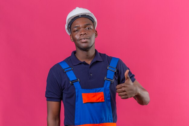 Joven constructor afroamericano hombre vestido con uniforme de construcción y casco de seguridad con una sonrisa en la cara mostrando el pulgar hacia arriba de pie en rosa