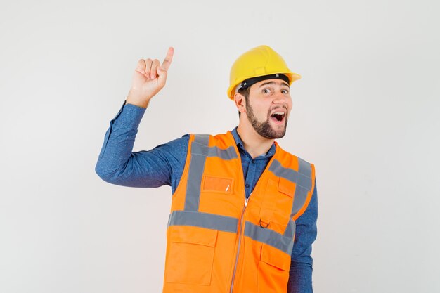 Joven constructor apuntando hacia arriba en camisa, chaleco, casco y mirando feliz. vista frontal.