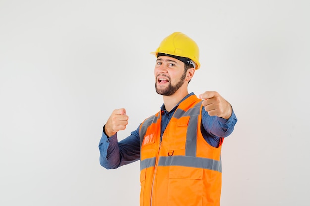 Joven constructor apuntando a la cámara en camisa, chaleco, casco y mirando confiado, vista frontal.