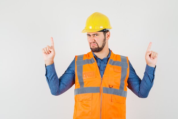 Joven constructor apuntando con el dedo hacia arriba en camisa, chaleco, casco y mirando triste, vista frontal.