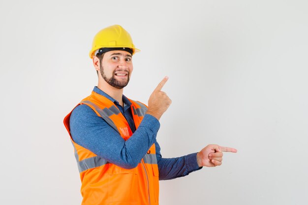 Joven constructor apuntando con el dedo hacia arriba y hacia un lado en camisa, chaleco, casco y mirando alegre. vista frontal.