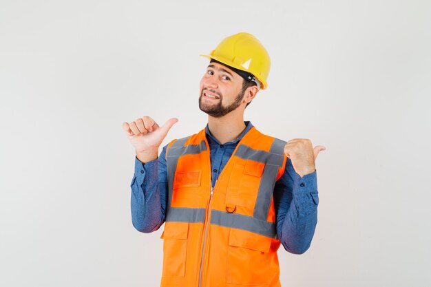 Joven constructor apuntando con los pulgares hacia atrás en camisa, chaleco, casco y mirando jovial. vista frontal.