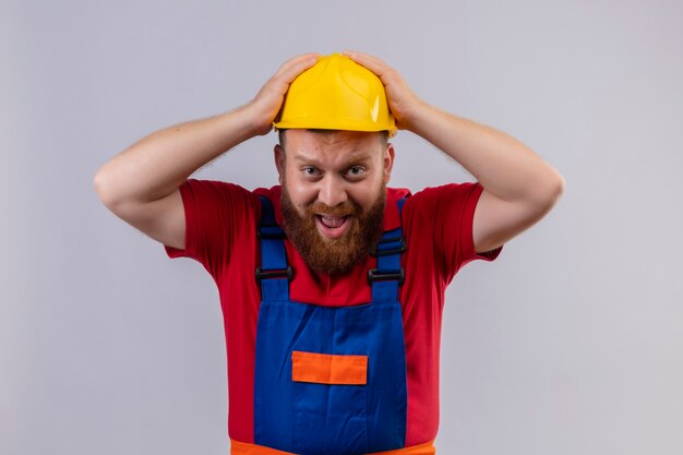 Joven constructor barbudo en uniforme de construcción y casco de seguridad mirando a cámara estresado y confundido tocando su cabeza gritando