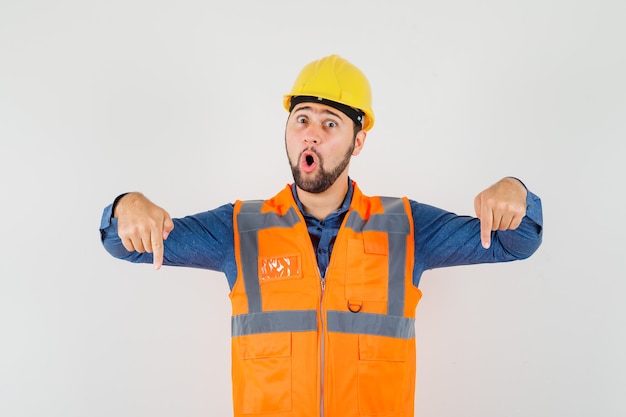 Joven constructor en camisa, chaleco, casco apuntando con el dedo hacia abajo y mirando asombrado, vista frontal.