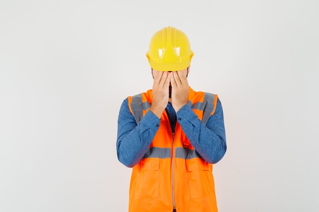 Joven constructor en camisa, chaleco, casco cubriendo la cara con las manos y mirando molesto, vista frontal.