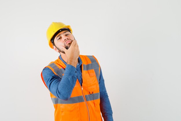 Joven constructor en camisa, chaleco, casco examinando la piel de la cara tocando su barba y luciendo guapo, vista frontal.