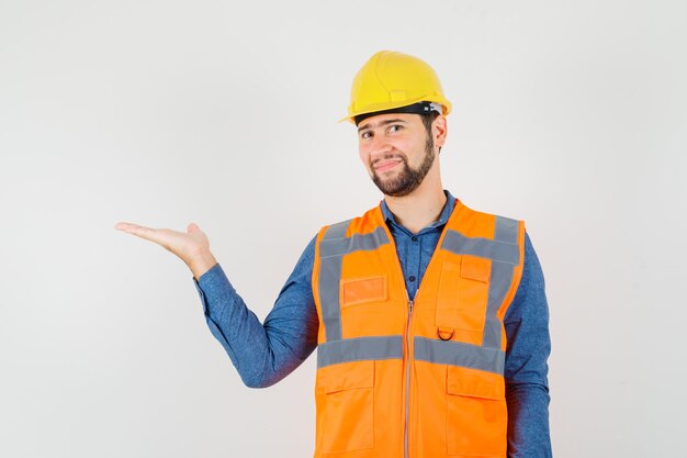 Joven constructor en camisa, chaleco, casco extendiendo la palma a un lado y mirando alegre, vista frontal.