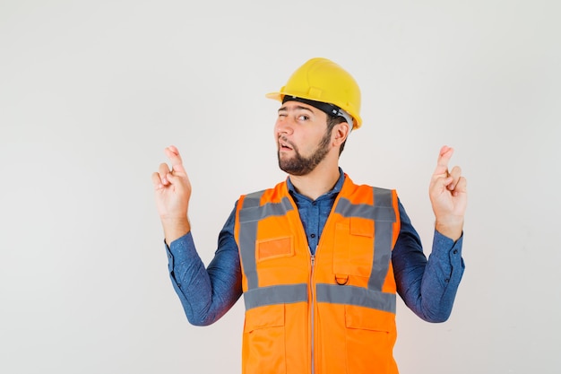 Joven constructor en camisa, chaleco, casco manteniendo los dedos cruzados y guiñando el ojo, vista frontal.