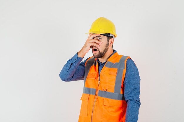 Joven constructor en camisa, chaleco, casco mirando a través de los dedos y mirando maravillado, vista frontal.