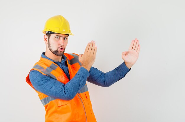 Joven constructor en camisa, chaleco, casco mostrando gesto de chuleta de karate y mirando confiado, vista frontal.