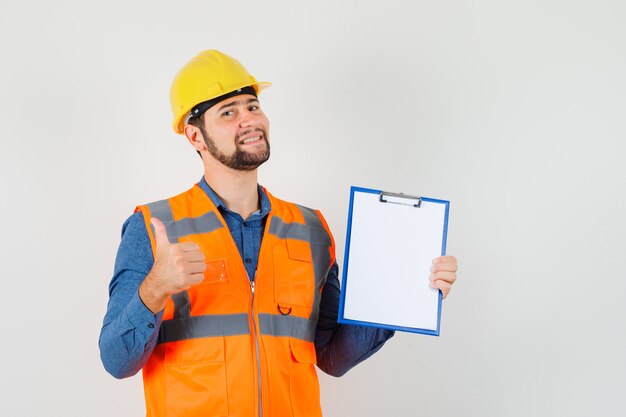 Joven constructor en camisa, chaleco, casco mostrando el pulgar hacia arriba, sosteniendo el portapapeles y mirando contento, vista frontal.