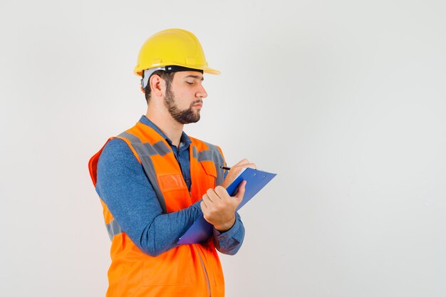 Joven constructor en camisa, chaleco, casco tomando notas en el portapapeles y mirando ocupado, vista frontal.