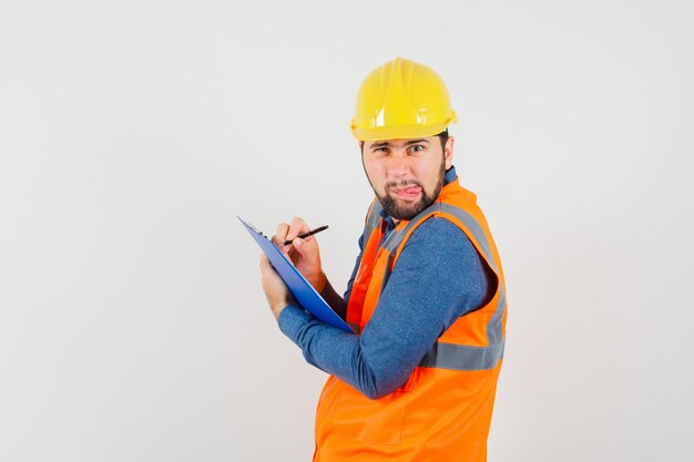 Joven constructor en camisa, chaleco, casco tomando notas en el portapapeles