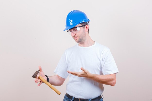 Joven constructor en camiseta, jeans, casco mostrando martillo y mirando pensativo