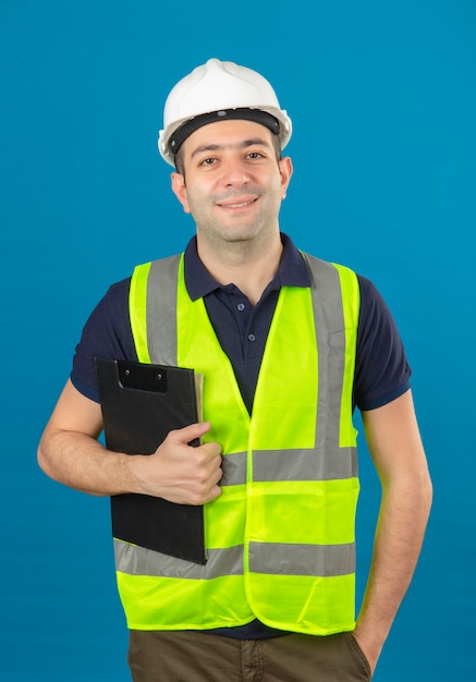 Joven constructor con casco blanco y un chaleco amarillo, sosteniendo el portapapeles con una sonrisa de pie en azul aislado